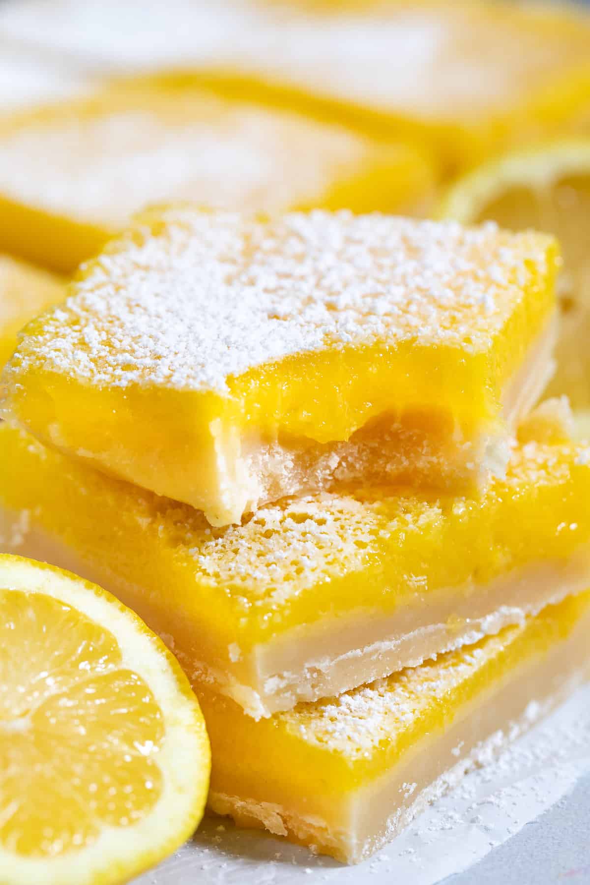 stacked lemon bars with powdered sugar on top around cut lemons.