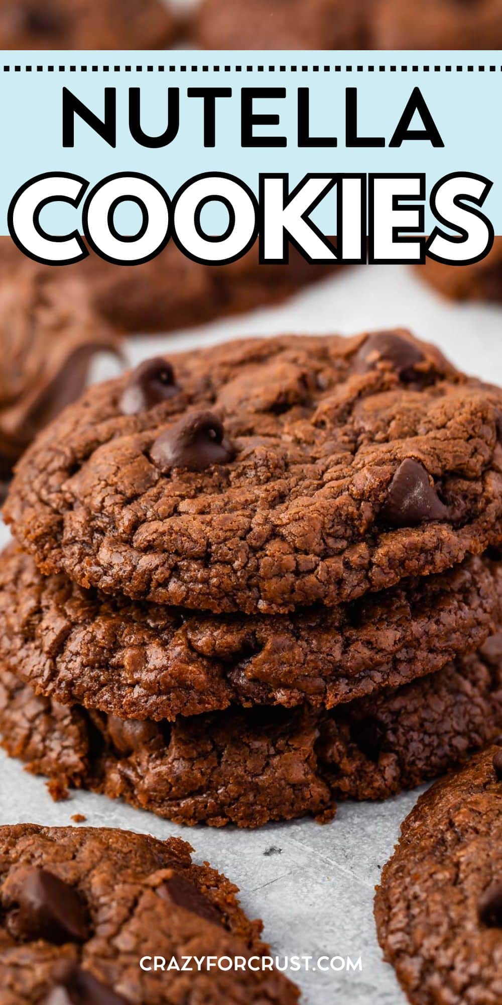 A stack of rich, chocolatey Nutella Cookies with melty chocolate chips sits on a light surface, with more cookies in the background. Text above reads Nutella Cookies and crazyforcrust.com is at the bottom.