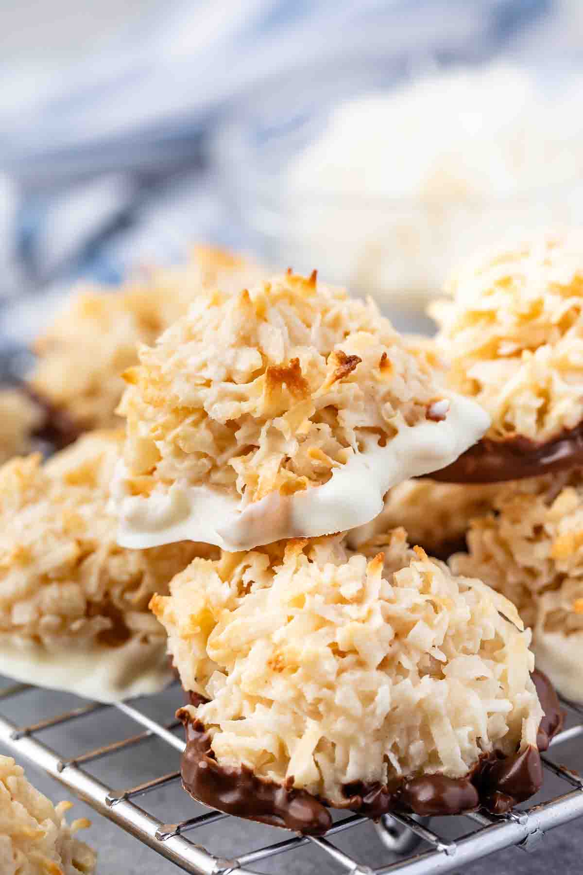 Close-up of macaroon cookies dipped in white and dark chocolate on a cooling rack. The golden-brown, shredded coconut tops contrast with the smooth, glossy chocolate bases. A blurred background adds depth to the image.