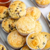 A plate of assorted pancake muffins—some dusted with powdered sugar, others studded with chocolate chips or berries—surrounded by a cup of syrup and a bowl of blueberries in the background.