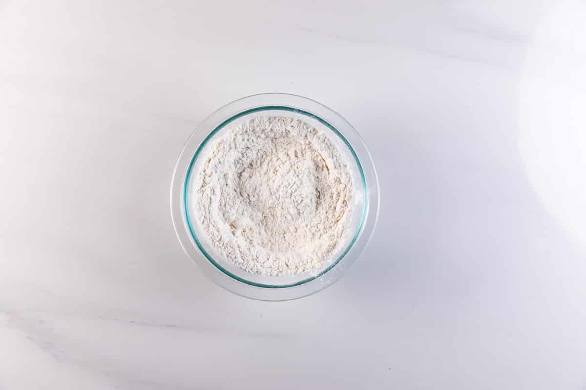 A glass bowl filled with white flour, perfect for making Pancake Muffins, sits on a white marble surface, viewed from above.