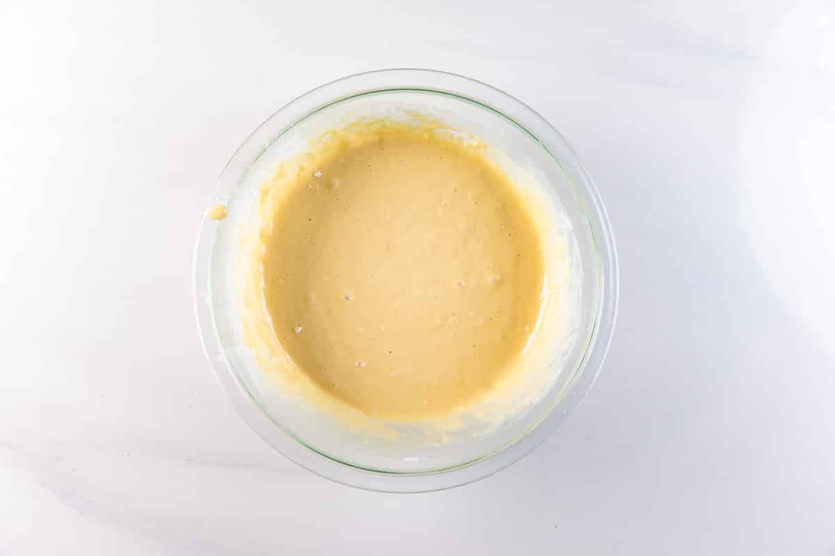 A glass bowl filled with smooth, pale yellow pancake muffins batter sits on a white marble surface.