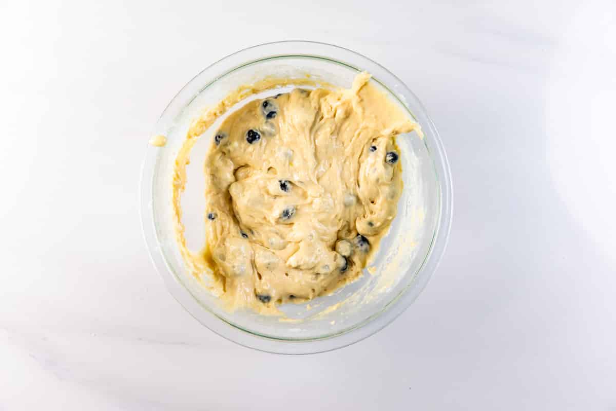 A glass bowl filled with thick, creamy pancake muffin batter mixed with blueberries, placed on a white marble surface.