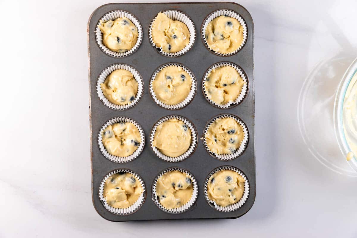 A muffin tray holds 12 paper liners filled with unbaked blueberry pancake muffin batter, ready for baking. The tray sits on a white surface, and a mixing bowl is partially visible on the right.