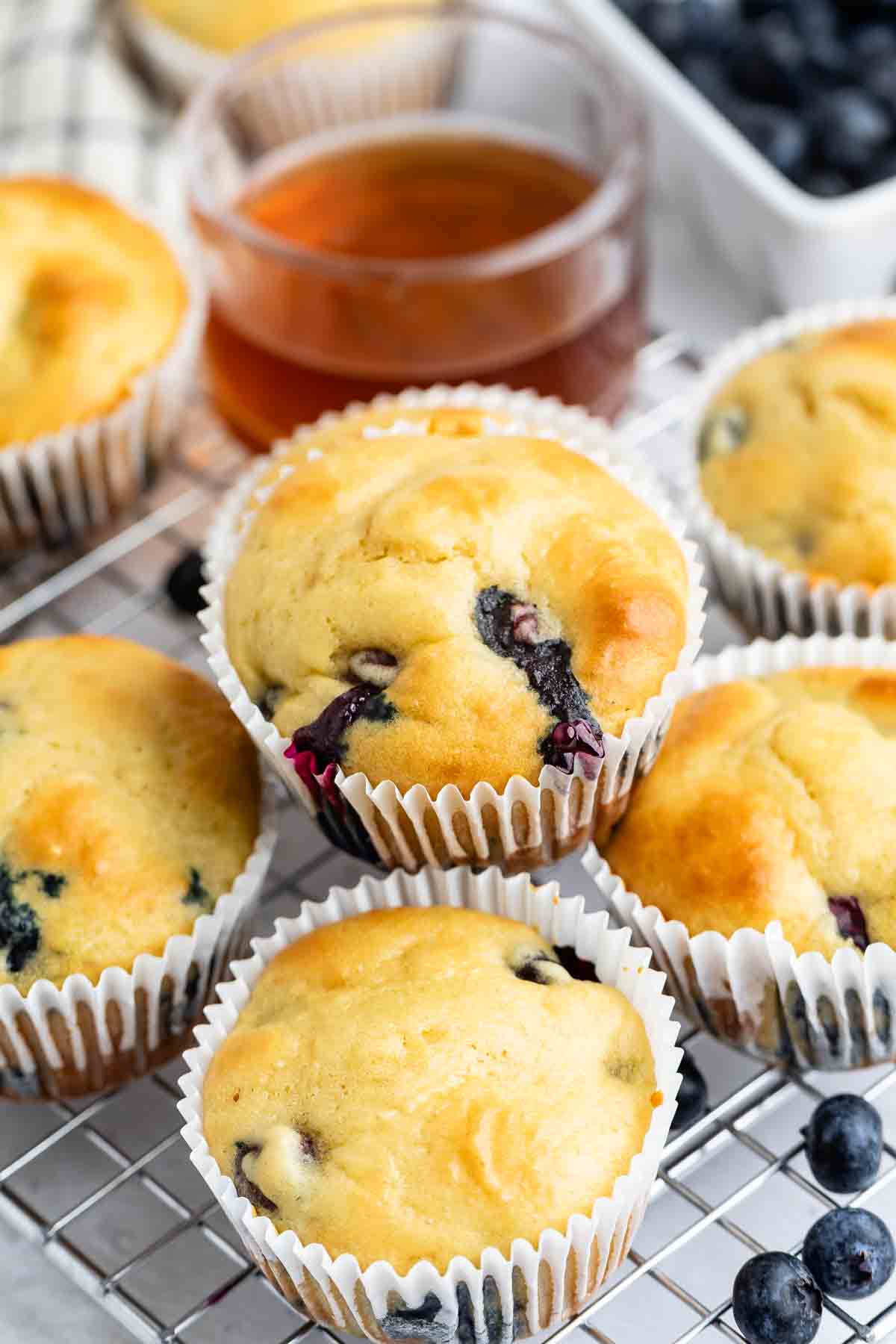 Freshly baked blueberry pancake muffins in white paper liners are cooling on a wire rack. A glass container of honey and a bowl of blueberries are in the background.