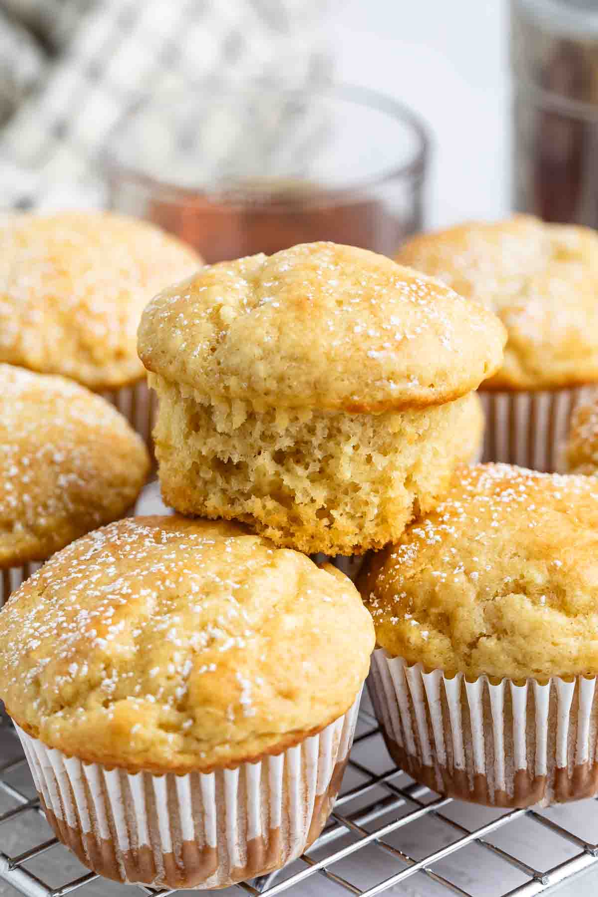 A close-up of golden brown Pancake Muffins topped with powdered sugar, arranged on a cooling rack. One muffin has a bite taken out, revealing its soft and moist interior.