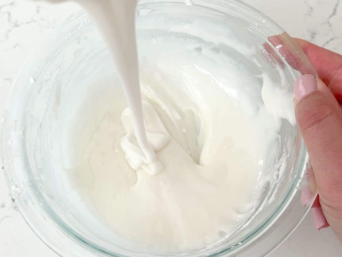 A hand holds a glass bowl while white sugar cookie icing is being poured and mixed inside. The mixture appears smooth and glossy on a white countertop.