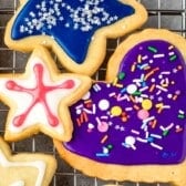 Three decorated sugar cookies—adorned with vibrant sugar cookie icing—rest on a cooling rack: a blue star sprinkled with white, a white star lined in red, and a purple heart topped with colorful sprinkles.