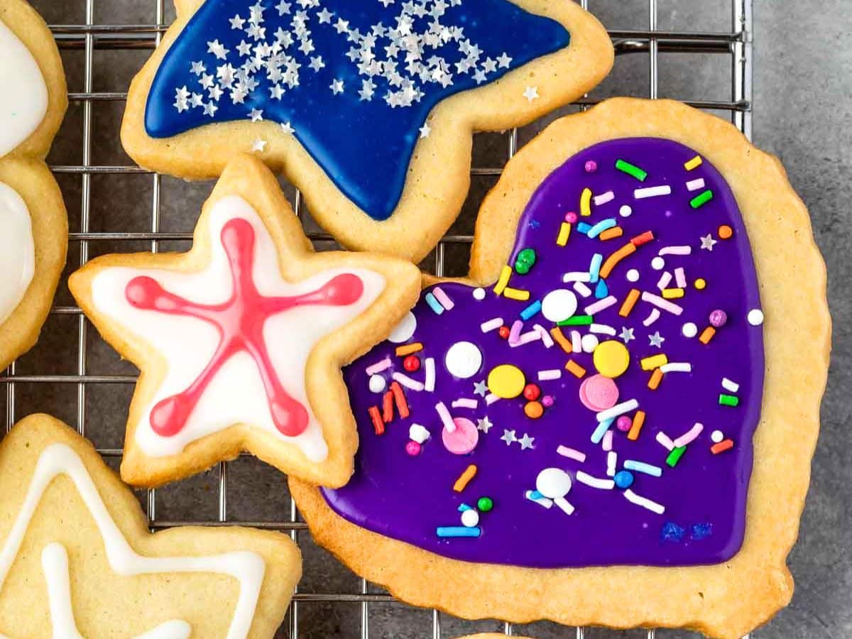 Three decorated sugar cookies—adorned with vibrant sugar cookie icing—rest on a cooling rack: a blue star sprinkled with white, a white star lined in red, and a purple heart topped with colorful sprinkles.