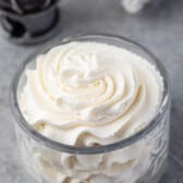 A glass bowl filled with swirled whipped cream sits on a gray surface, hinting at a perfect whipped cream recipe, with a blurred whisk and piping tip in the background.