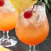 orange cocktail in a clear glass with a cherry and pineapple slice on the rim.