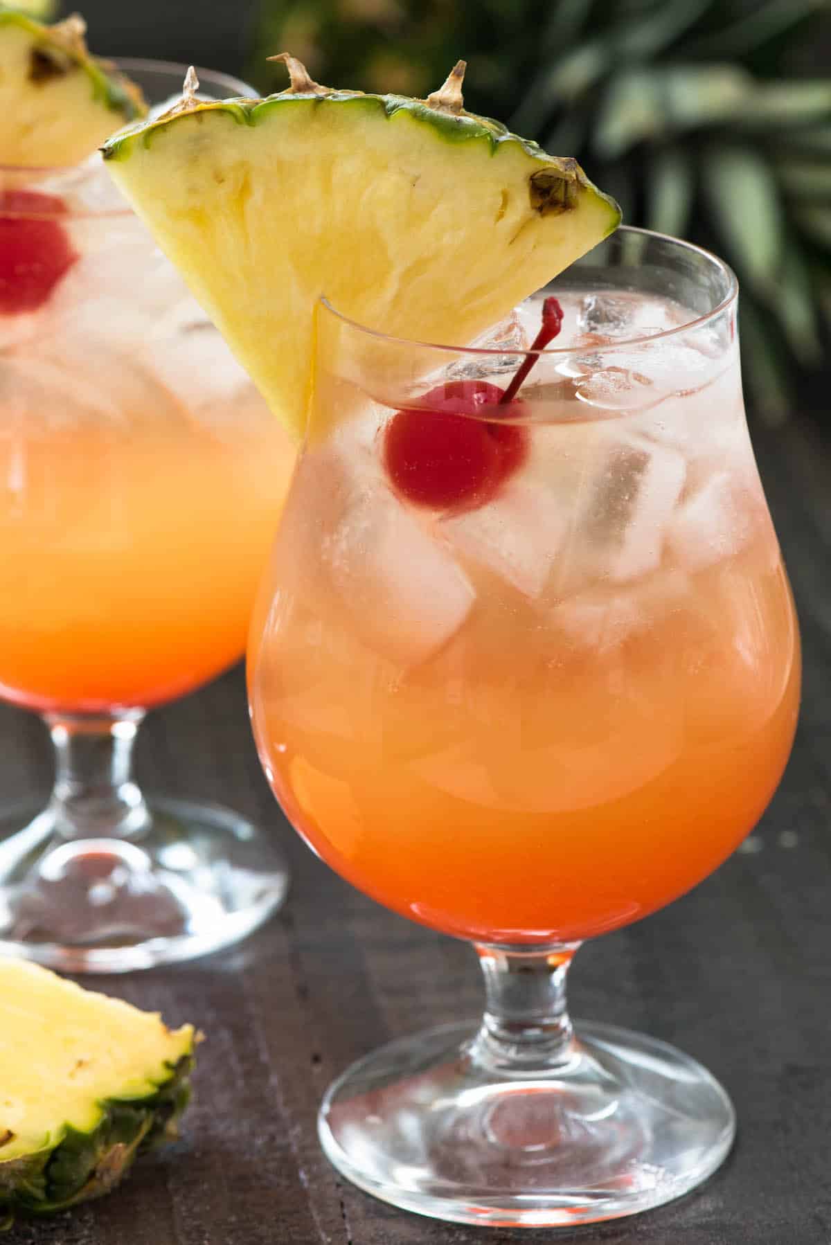 orange cocktail in a clear glass with a cherry and pineapple slice on the rim.