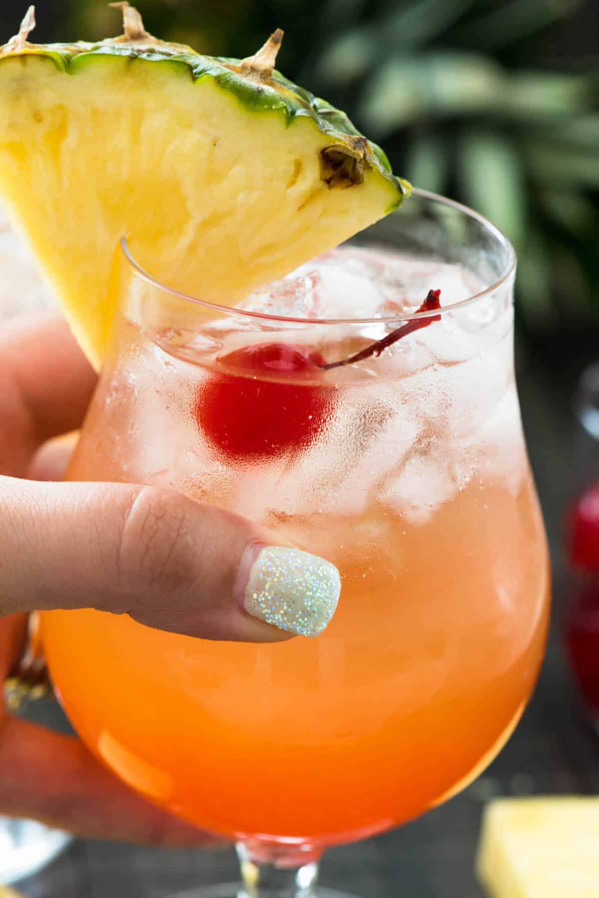 orange cocktail in a clear glass with a cherry and pineapple slice on the rim.