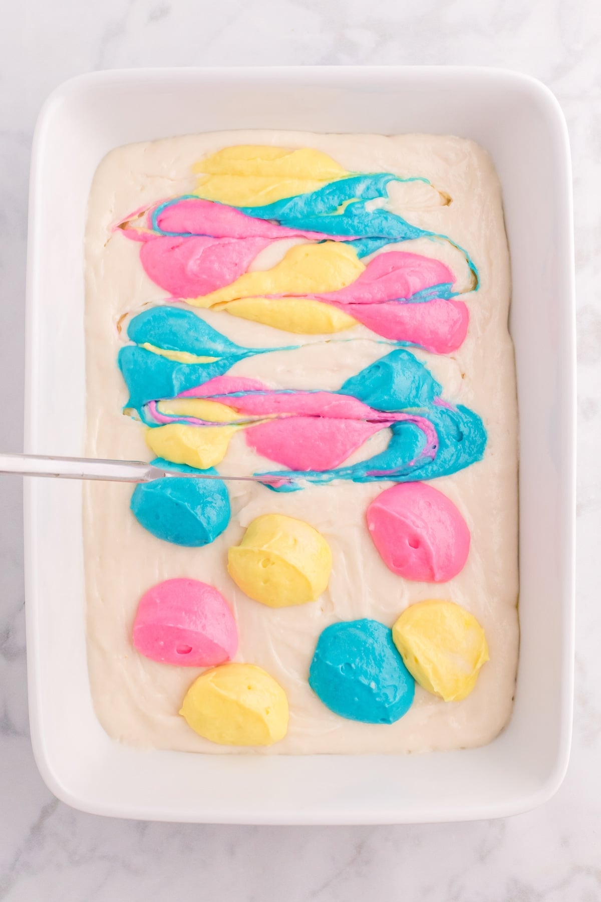 A baking dish filled with tie dye cake batter, with dollops of bright pink, yellow, and blue swirled together using a metal utensil to create a marbled, colorful pattern on top.