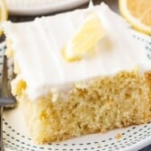 A slice of Lemon Cake with white icing and a small lemon wedge on top sits on a white plate with blue dots, next to a fork and fresh lemon slices in the background.