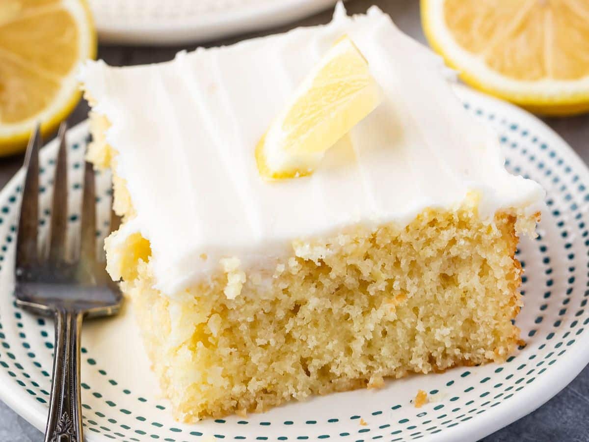 A slice of Lemon Cake with white icing and a small lemon wedge on top sits on a white plate with blue dots, next to a fork and fresh lemon slices in the background.