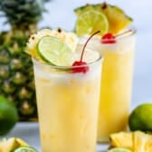 yellow cocktail in a clear glass with a cherry, lime and pineapple slice in the drink.