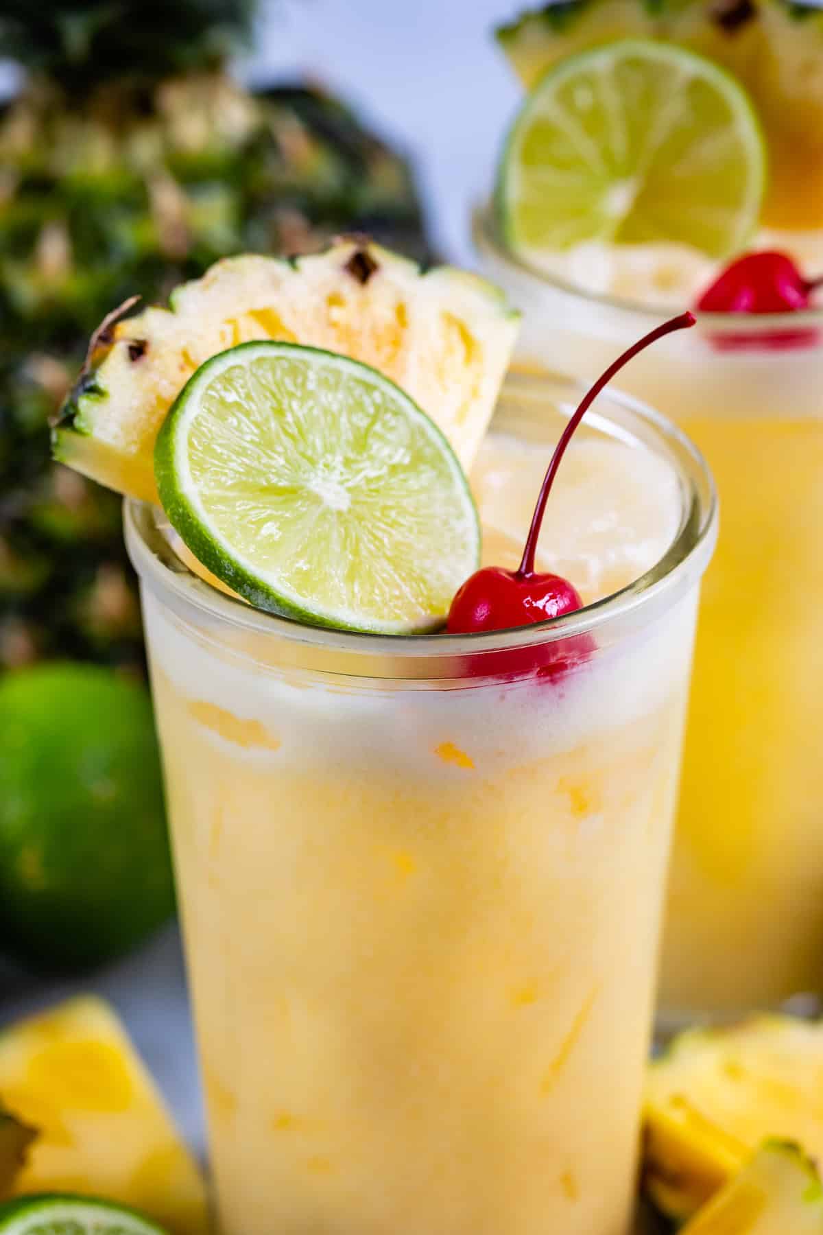 yellow cocktail in a clear glass with a cherry, lime and pineapple slice in the drink.