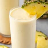 yellow pineapple smoothie in a tall clear glass with a pineapple slice on the rim.