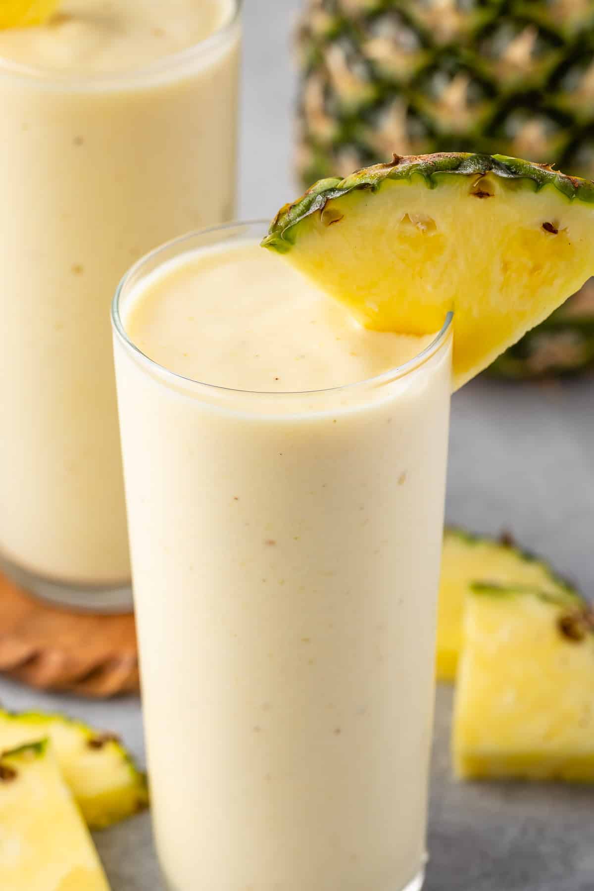 yellow pineapple smoothie in a tall clear glass with a pineapple slice on the rim.