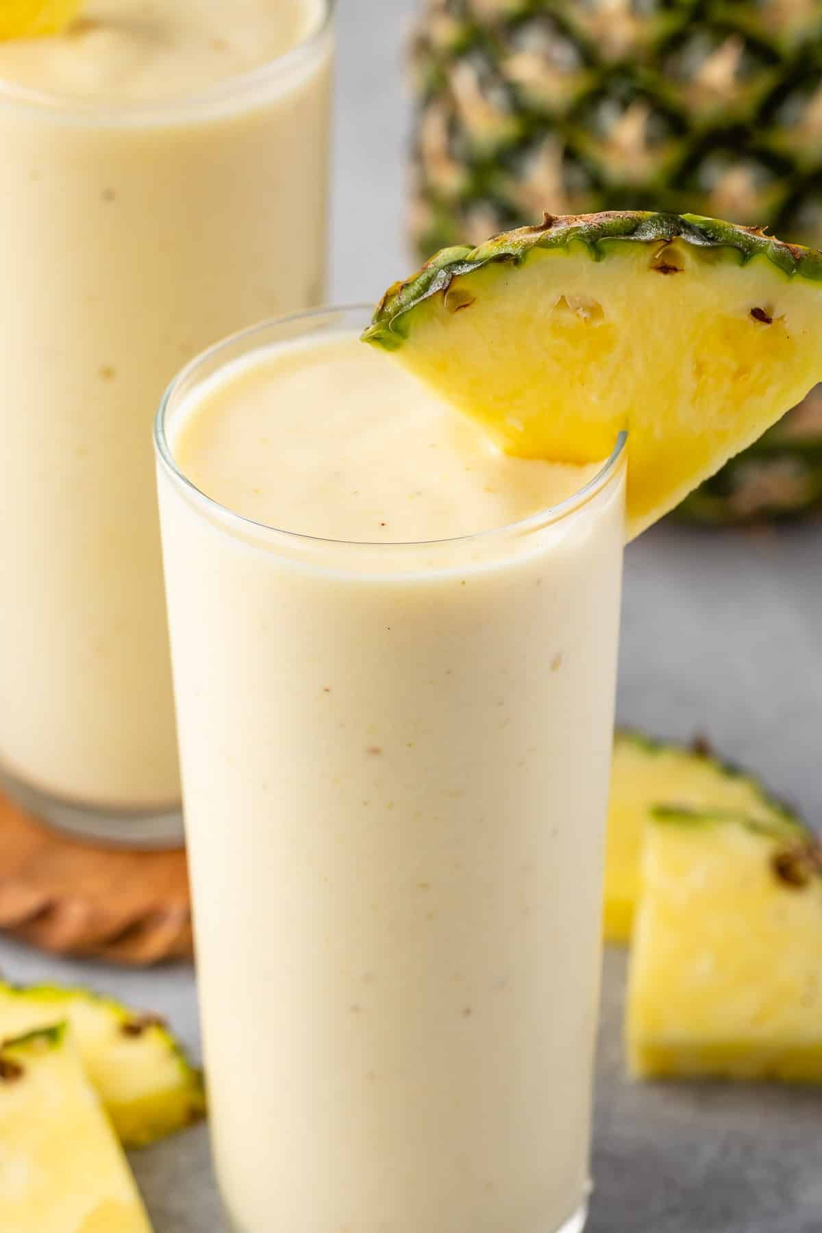yellow pineapple smoothie in a tall clear glass with a pineapple slice on the rim.