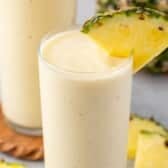 yellow pineapple smoothie in a tall clear glass with a pineapple slice on the rim with words on the image.