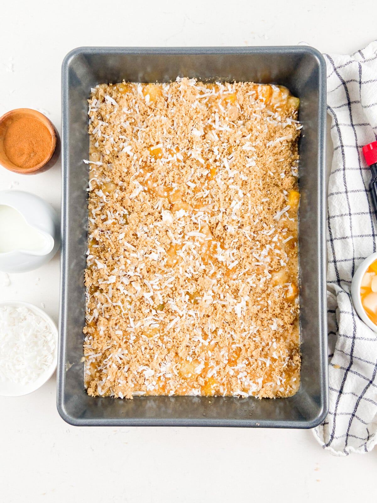 A baking pan filled with unbaked Fruit Cocktail Cake is topped with shredded coconut and a crumb mixture. Surrounding the pan are a bowl of cinnamon, a jug of milk, a bowl of coconut, and a cloth towel.