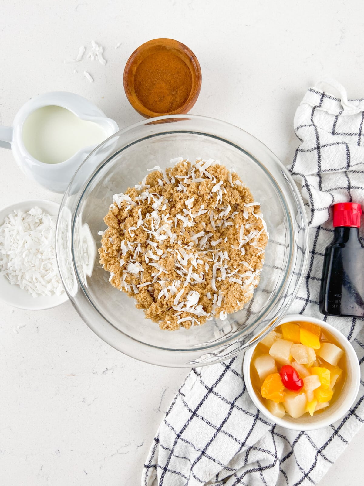 A glass bowl filled with a crumbly mixture and shredded coconut sits surrounded by milk, a wooden bowl, a dish of fruit cocktail, vanilla, more coconut, and a checkered towel—perfect for assembling Fruit Cocktail Cake.