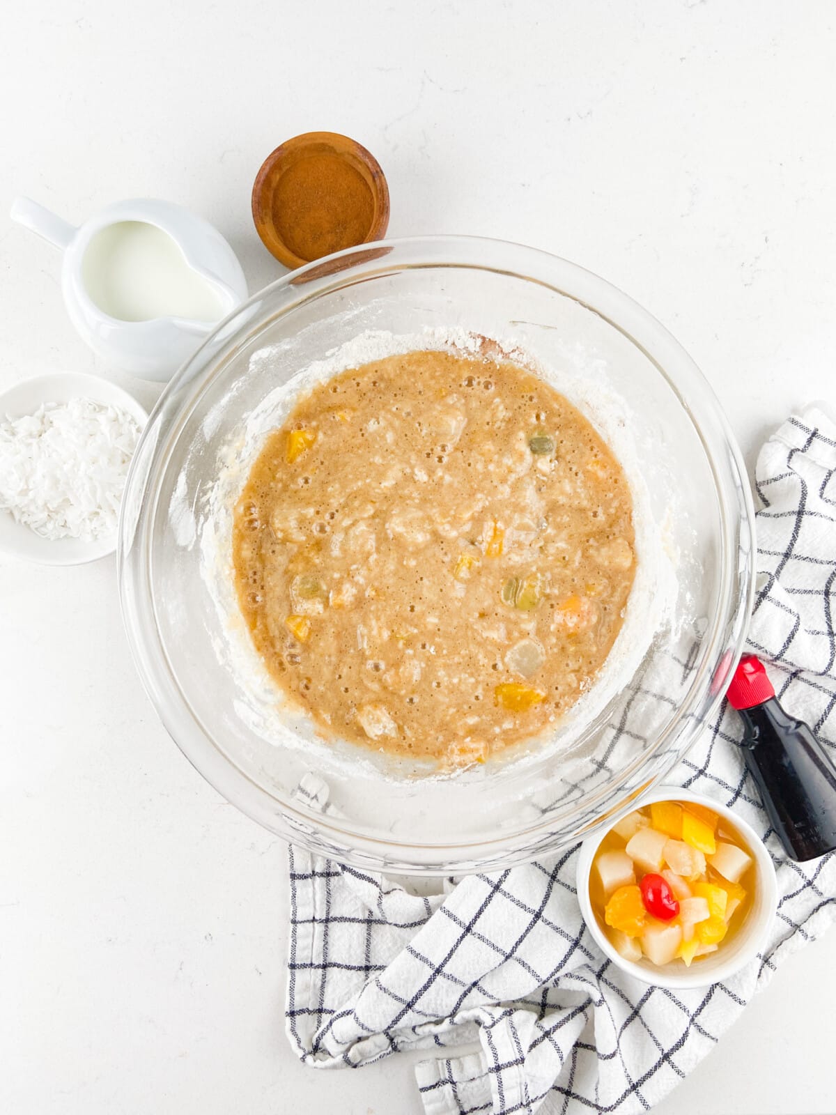 A glass bowl with mixed batter for Fruit Cocktail Cake, containing fruit pieces, sits on a white surface, surrounded by a bowl of mixed fruit, shredded coconut, a small pitcher of milk, a wooden bowl, and a checkered kitchen towel.