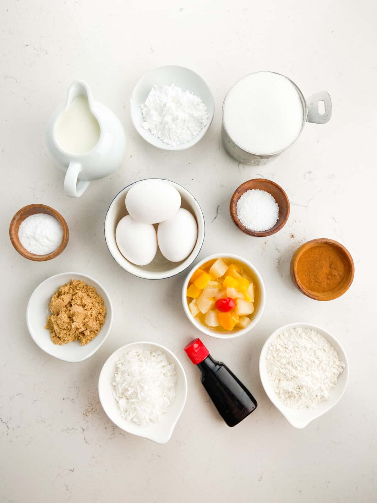 Top-down view of baking ingredients for Fruit Cocktail Cake arranged in bowls and measuring cups on a white surface, including eggs, milk, flour, sugar, vanilla extract, cinnamon, coconut, and canned fruit salad.