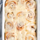 A white baking dish filled with freshly baked overnight cinnamon rolls, covered in a thick layer of creamy icing, seen from above on a light gray countertop.