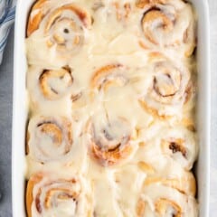 A white baking dish filled with freshly baked overnight cinnamon rolls, covered in a thick layer of creamy icing, seen from above on a light gray countertop.