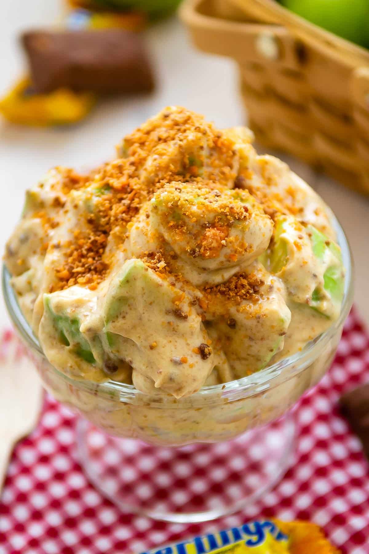 apple salad in a clear raised bowl covered with crushed butterfinger candy bar.