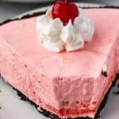 A slice of pink creamy jello pie with a chocolate cookie crust, topped with a swirl of whipped cream and a maraschino cherry, on a white plate.