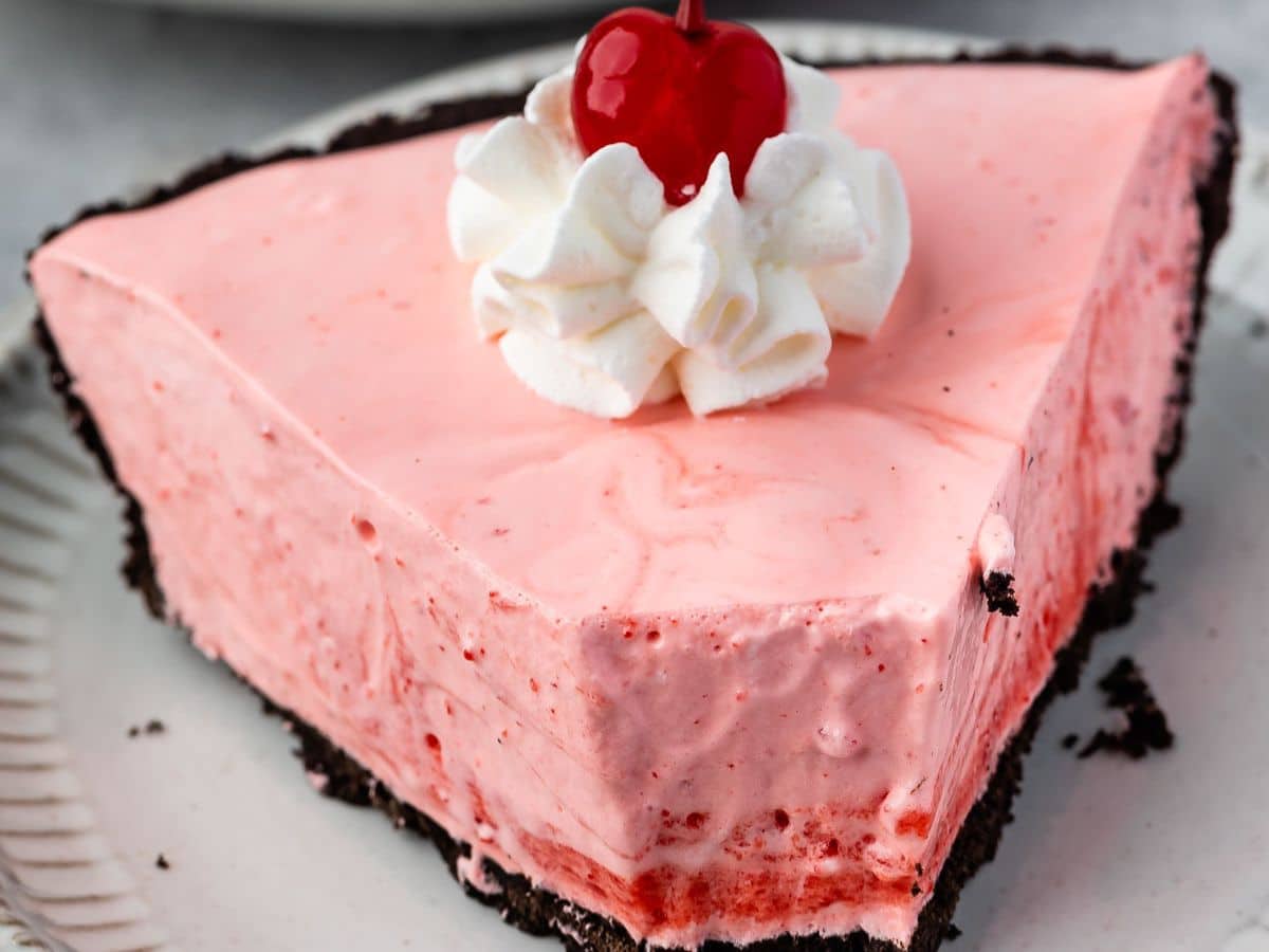 A slice of pink creamy jello pie with a chocolate cookie crust, topped with a swirl of whipped cream and a maraschino cherry, on a white plate.