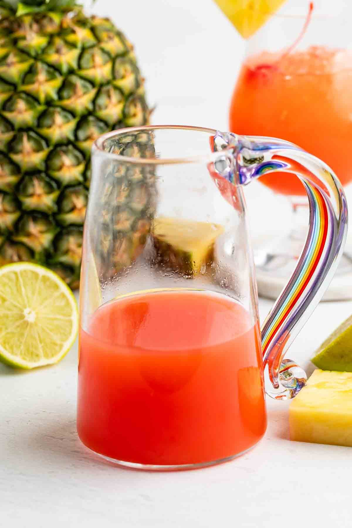 A glass pitcher with a colorful handle contains red juice, perfect for serving your favorite rum punch recipe. Nearby are a whole pineapple, a halved lime, pineapple slices, and a glass of red drink garnished with pineapple.