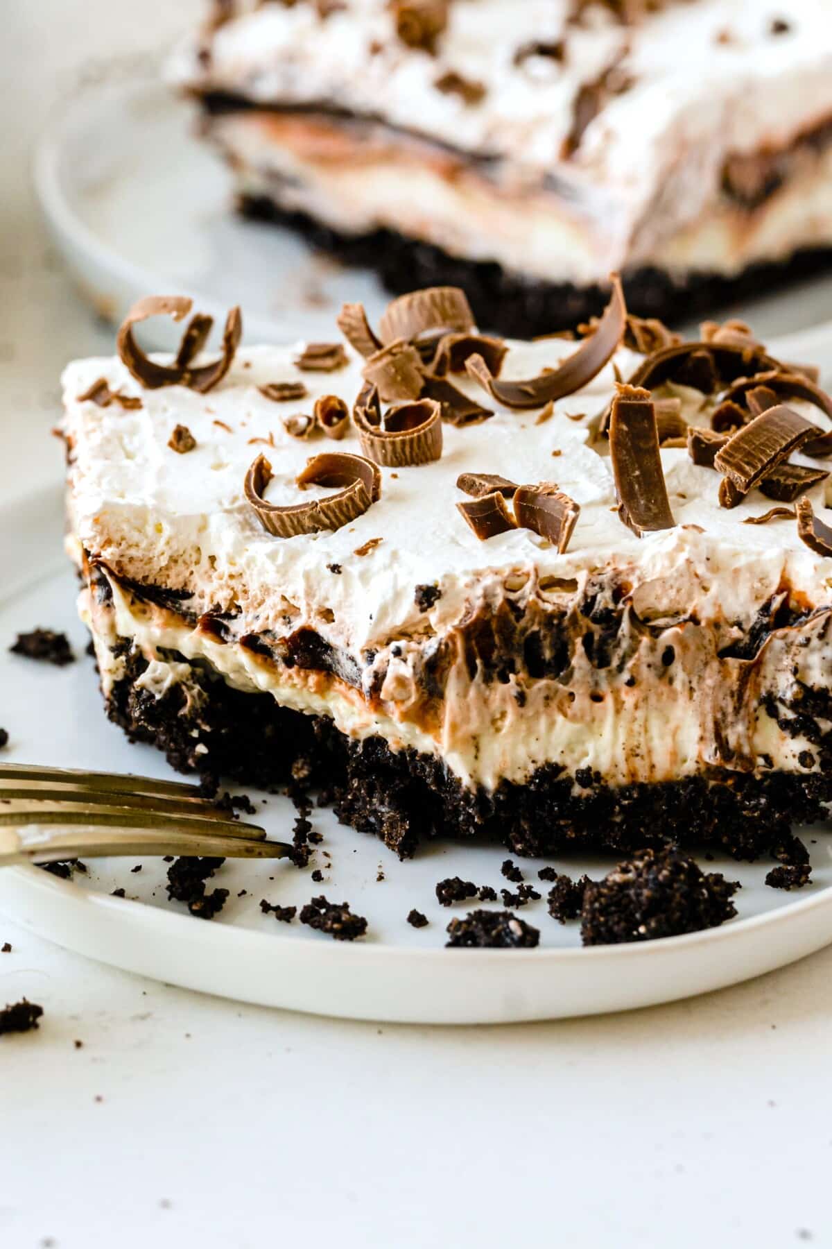A slice of chocolate lasagna with a cookie crust, creamy filling, whipped topping, and chocolate curls is served on a white plate, with a fork and crumbs beside it.