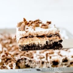 A close-up of a layered chocolate lasagna dessert bar with whipped topping, chocolate shavings, and peanut butter cups, being lifted from a baking pan.