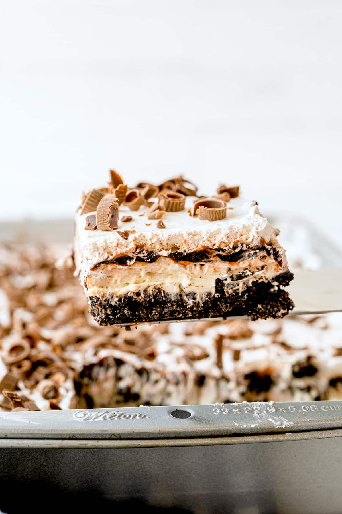 A close-up of a layered chocolate lasagna dessert bar with whipped topping, chocolate shavings, and peanut butter cups, being lifted from a baking pan.