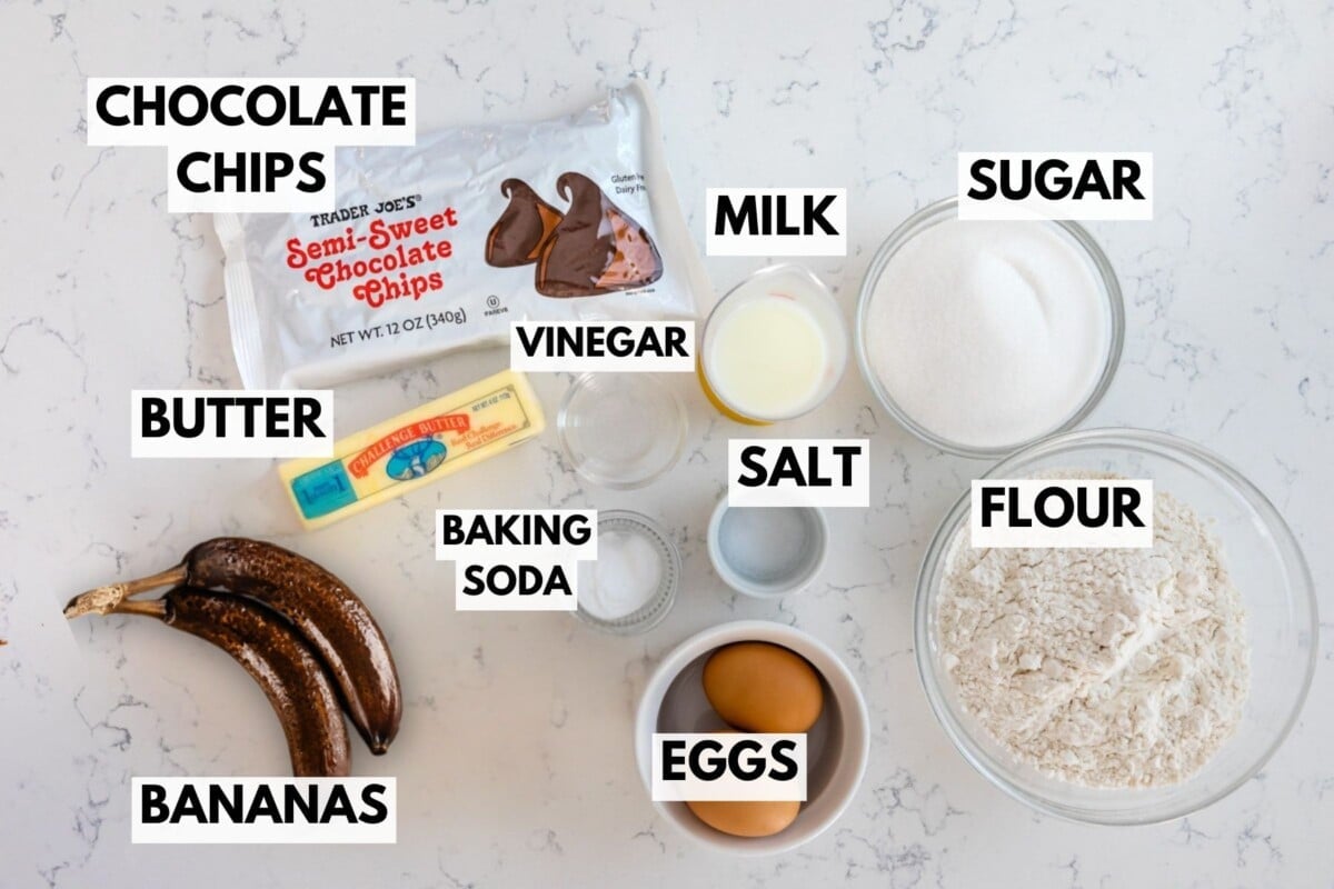 A flat lay of Chocolate Chip Banana Bread ingredients&mdash;chocolate chips, butter, ripe bananas, baking soda, vinegar, milk, eggs, salt, flour, and sugar&mdash;neatly arranged and labeled on a white marble surface.