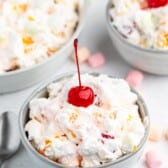 A bowl of creamy Tropical Fluff fruit salad topped with a bright red cherry, surrounded by colorful mini marshmallows and two more bowls of the same salad in the background. A spoon rests beside the bowl on a white surface.