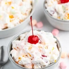 A bowl of creamy Tropical Fluff fruit salad topped with a bright red cherry, surrounded by colorful mini marshmallows and two more bowls of the same salad in the background. A spoon rests beside the bowl on a white surface.
