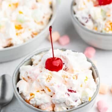 A bowl of creamy Tropical Fluff fruit salad topped with a bright red cherry, surrounded by colorful mini marshmallows and two more bowls of the same salad in the background. A spoon rests beside the bowl on a white surface.