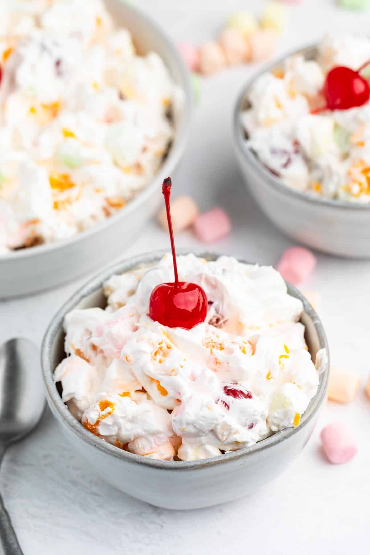 A bowl of creamy Tropical Fluff fruit salad topped with a bright red cherry, surrounded by colorful mini marshmallows and two more bowls of the same salad in the background. A spoon rests beside the bowl on a white surface.