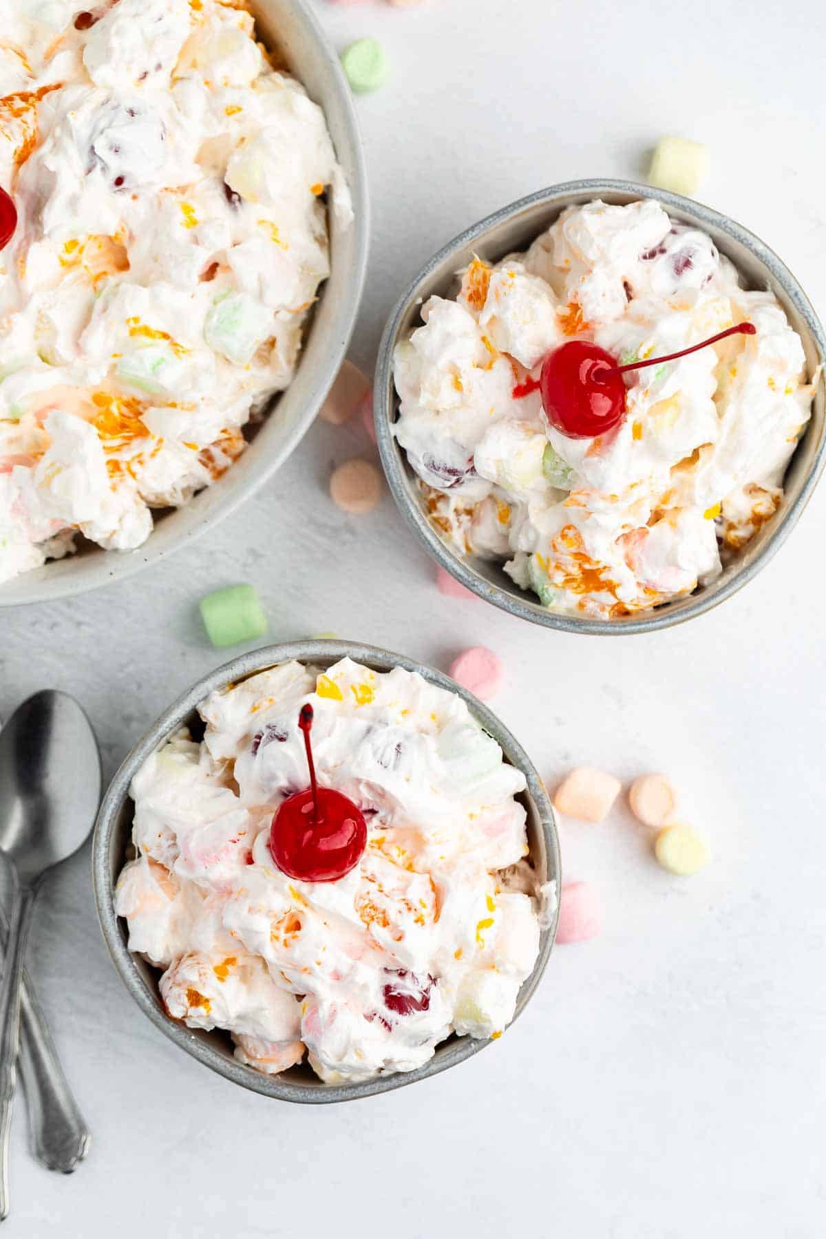 Three bowls of creamy Tropical Fluff fruit salad topped with maraschino cherries, surrounded by pastel mini marshmallows on a light surface, with two spoons placed nearby.