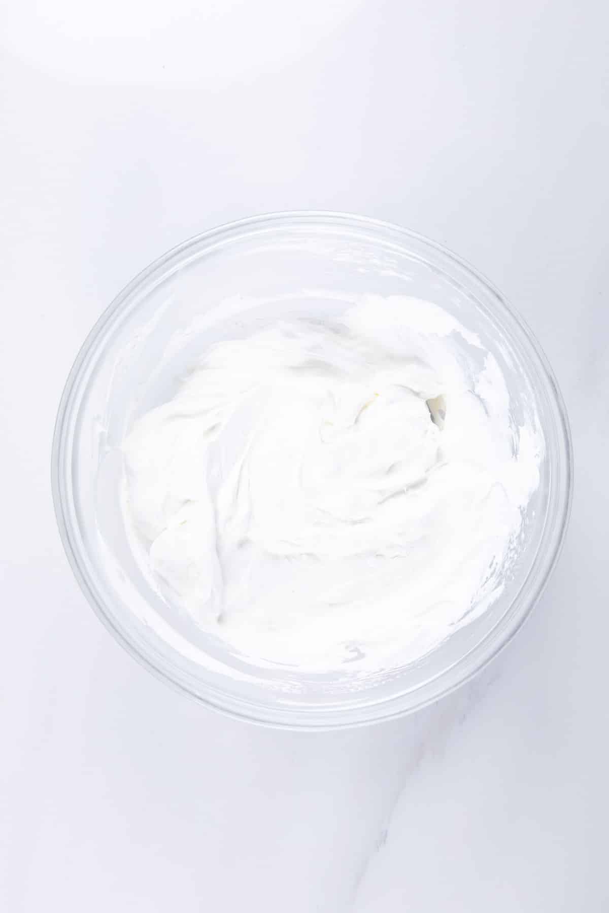 A clear glass bowl filled with smooth, white Tropical Fluff whipped cream or frosting sits on a white marble surface.