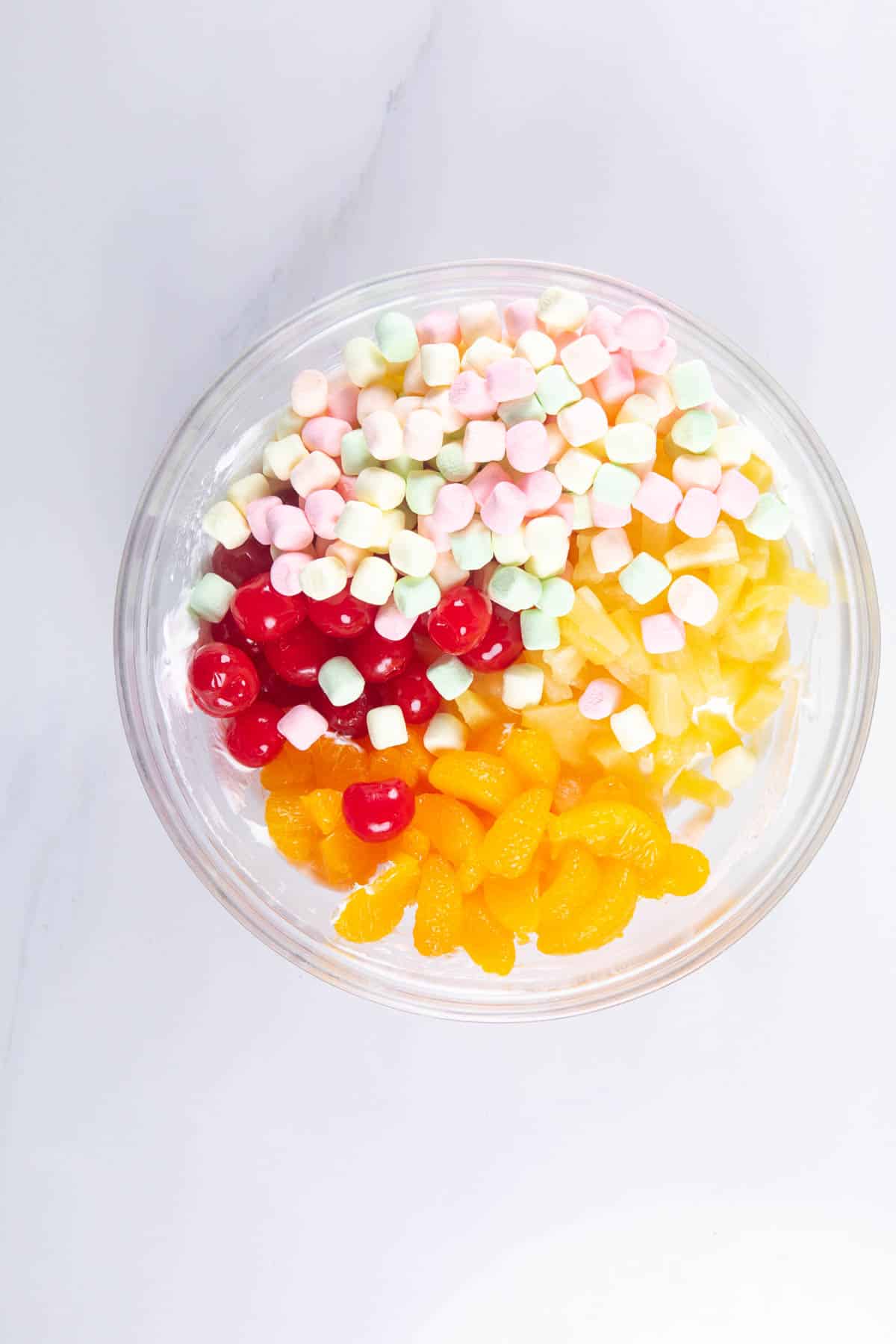 A glass bowl filled with colorful mini marshmallows, red cherries, pineapple chunks, and mandarin orange segments—each arranged in separate sections—creates a vibrant Tropical Fluff display on a white surface.