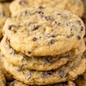 A close-up of a stack of banana chocolate chip cookies, showing their golden-brown color and visible chocolate chips, with more cookies blurred in the background.
