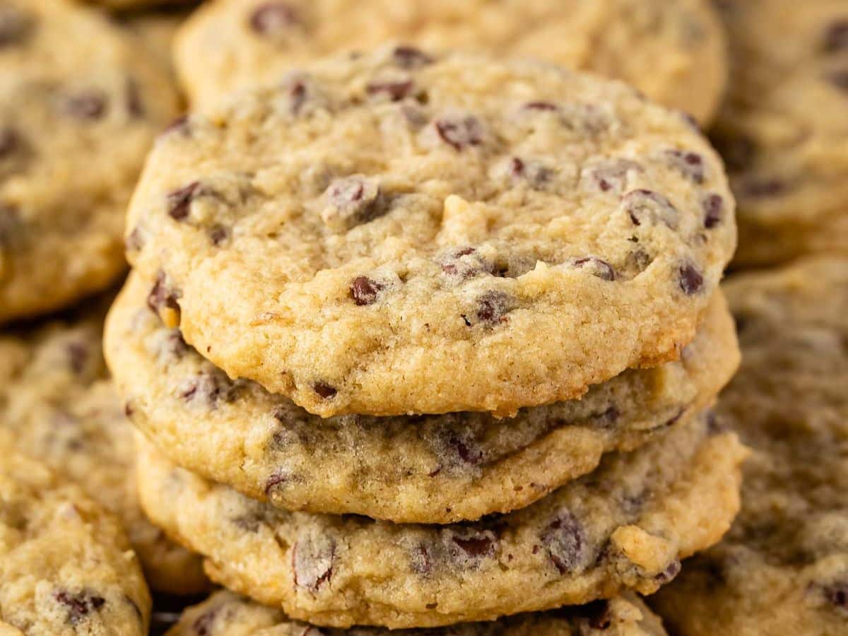 A close-up of a stack of banana chocolate chip cookies, showing their golden-brown color and visible chocolate chips, with more cookies blurred in the background.