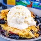 A scoop of vanilla ice cream sits on top of a golden blueberry dump cake cobbler, garnished with fresh blueberries and served on a gray plate.
